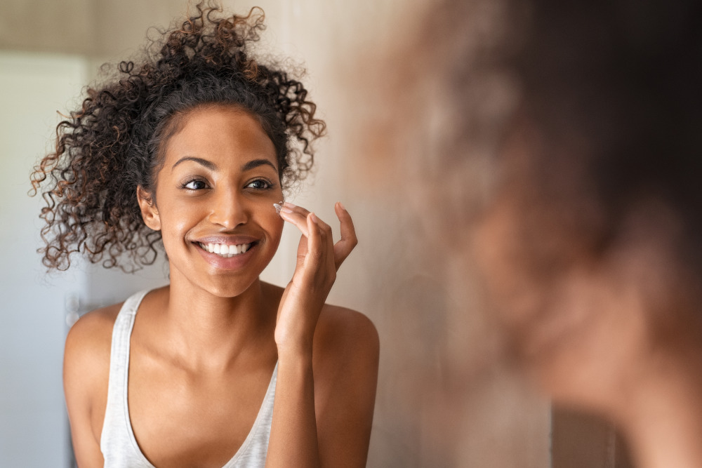 Young woman in while tank top looking in the mirror while applying sunscreen to avoid getting sunspots.