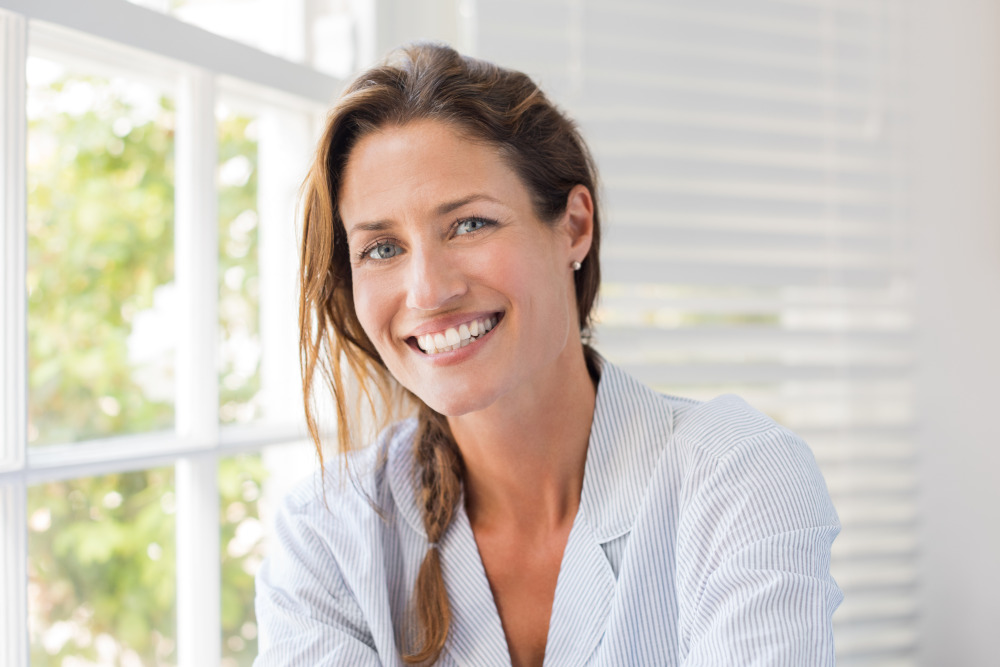 Smiling woman in a blue pinstriped blazer sitting in front of her window.